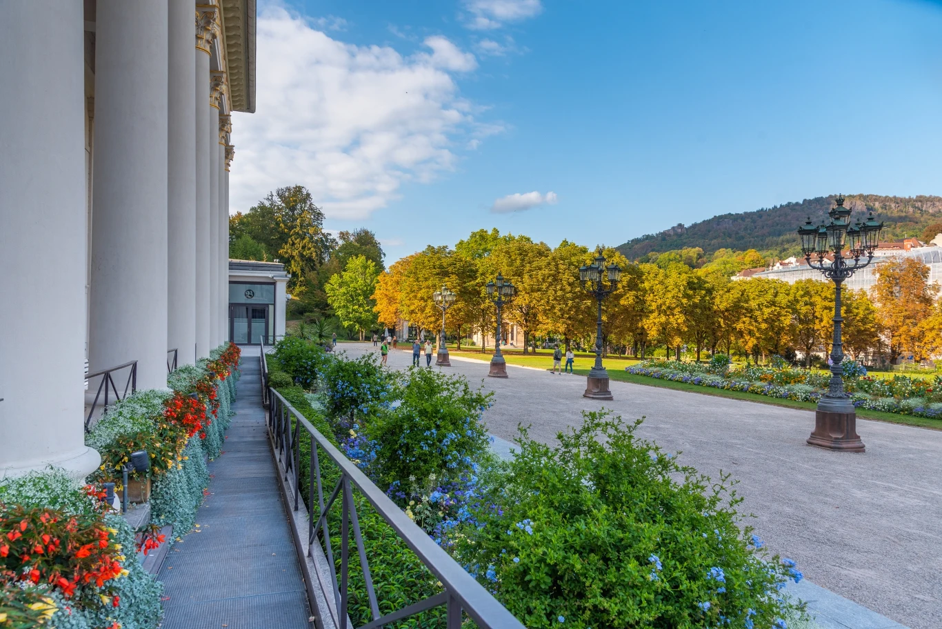 Szeroka promenada ozdobiona eleganckimi lampami ulicznymi, otoczona kolorowymi kwiatami i drzewami, z monumentalnym budynkiem po lewej stronie oraz widokiem na zielone wzgórza w tle.