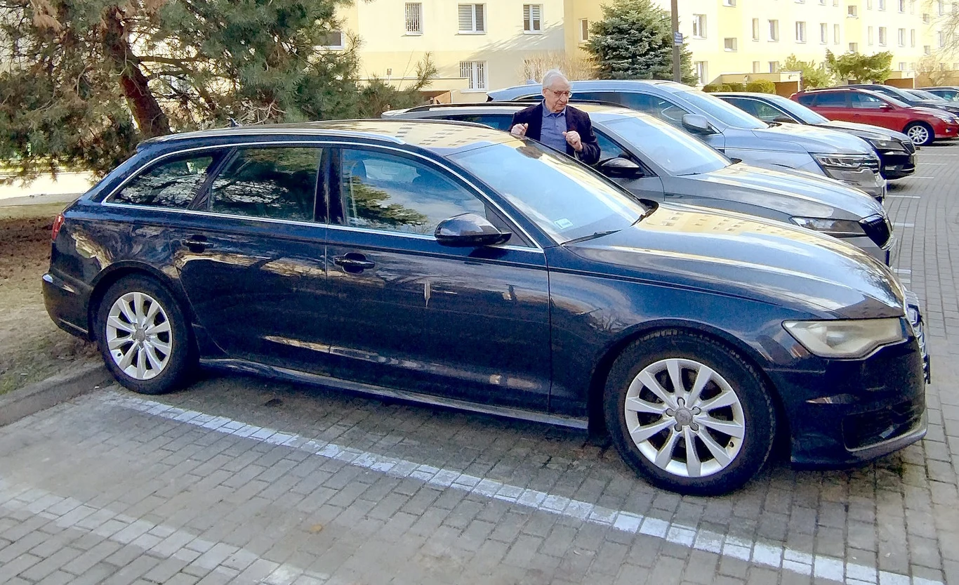 Ciemny kombi zaparkowany na parkingu obok innych samochodów, mężczyzna stoi przy sąsiadującym aucie trzymając kluczyki.