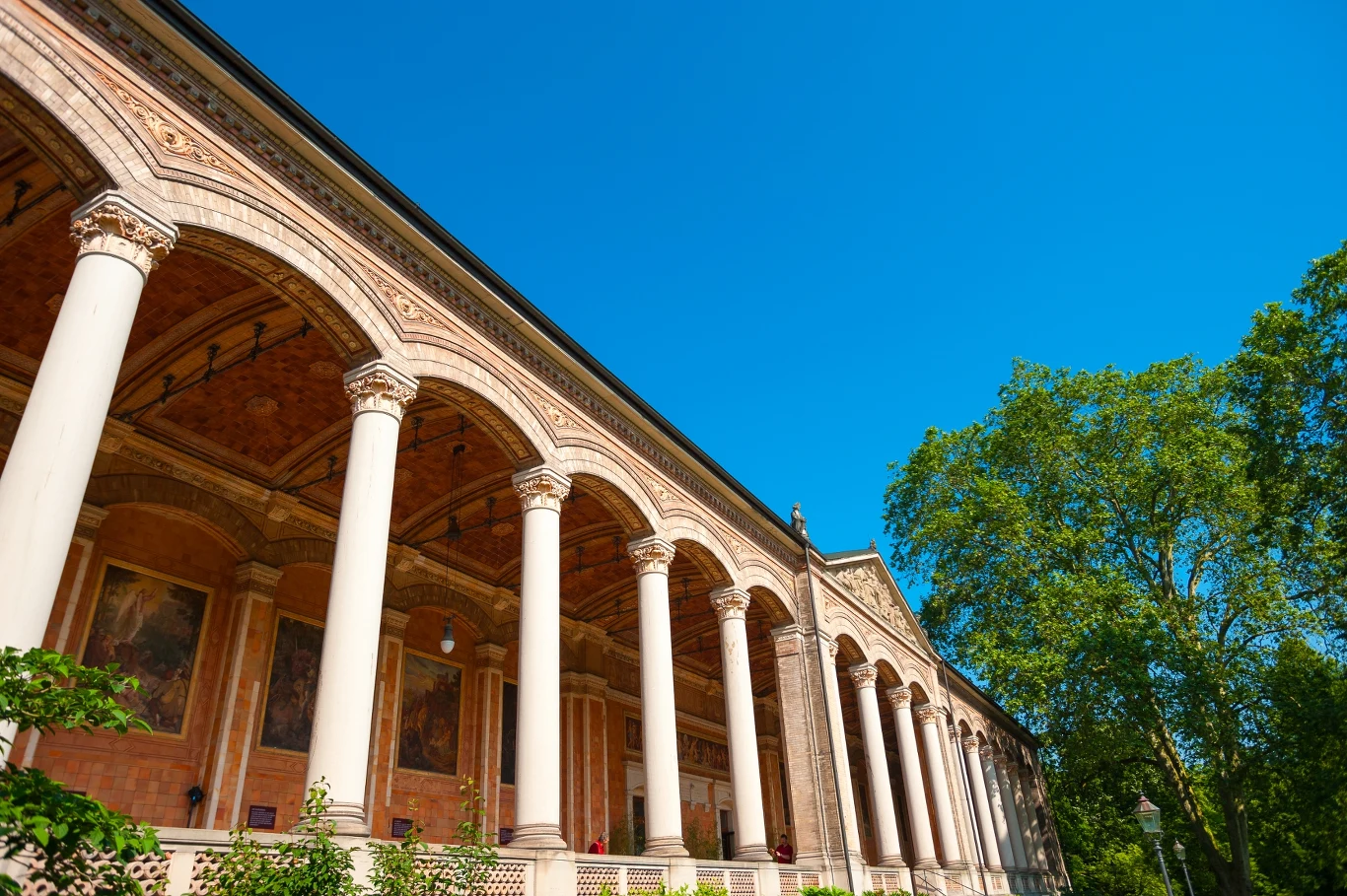 Rząd wysokich, białych kolumn wspierających zdobiony gzyms budynku z freskami pod arkadami, otoczony zielenią drzew na tle jasnobłękitnego nieba.