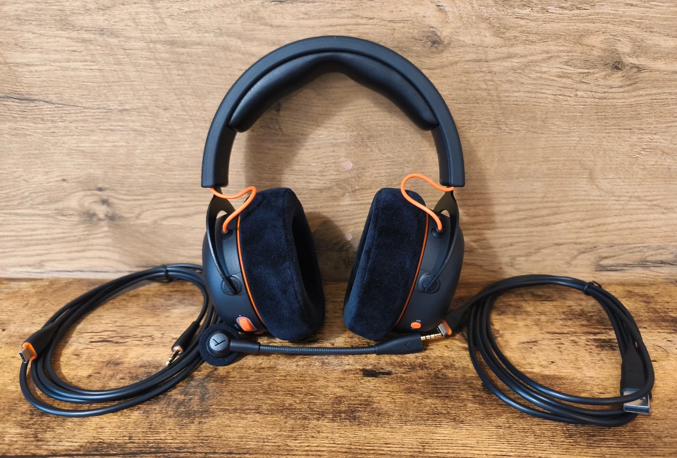 Beyerdynamic MMX 150 Wireless Czarne nauszne słuchawki z miękkimi, welurowymi poduszkami, pomarańczowymi akcentami przy nausznicach oraz mikrofonem na pałąku, ułożone na drewnianej powierzchni wraz z dwoma spiralnie zwiniętymi kablami po bokach.