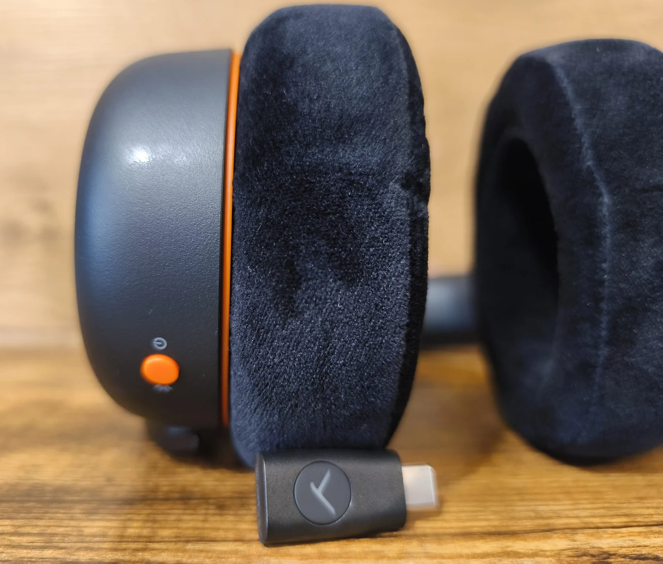 Beyerdynamic MMX 150 Wireless Czarne, welurowe nauszniki słuchawek z widocznym pomarańczowym przyciskiem i małym portem USB-C, leżące na drewnianej powierzchni obok adaptera USB-C z logo.