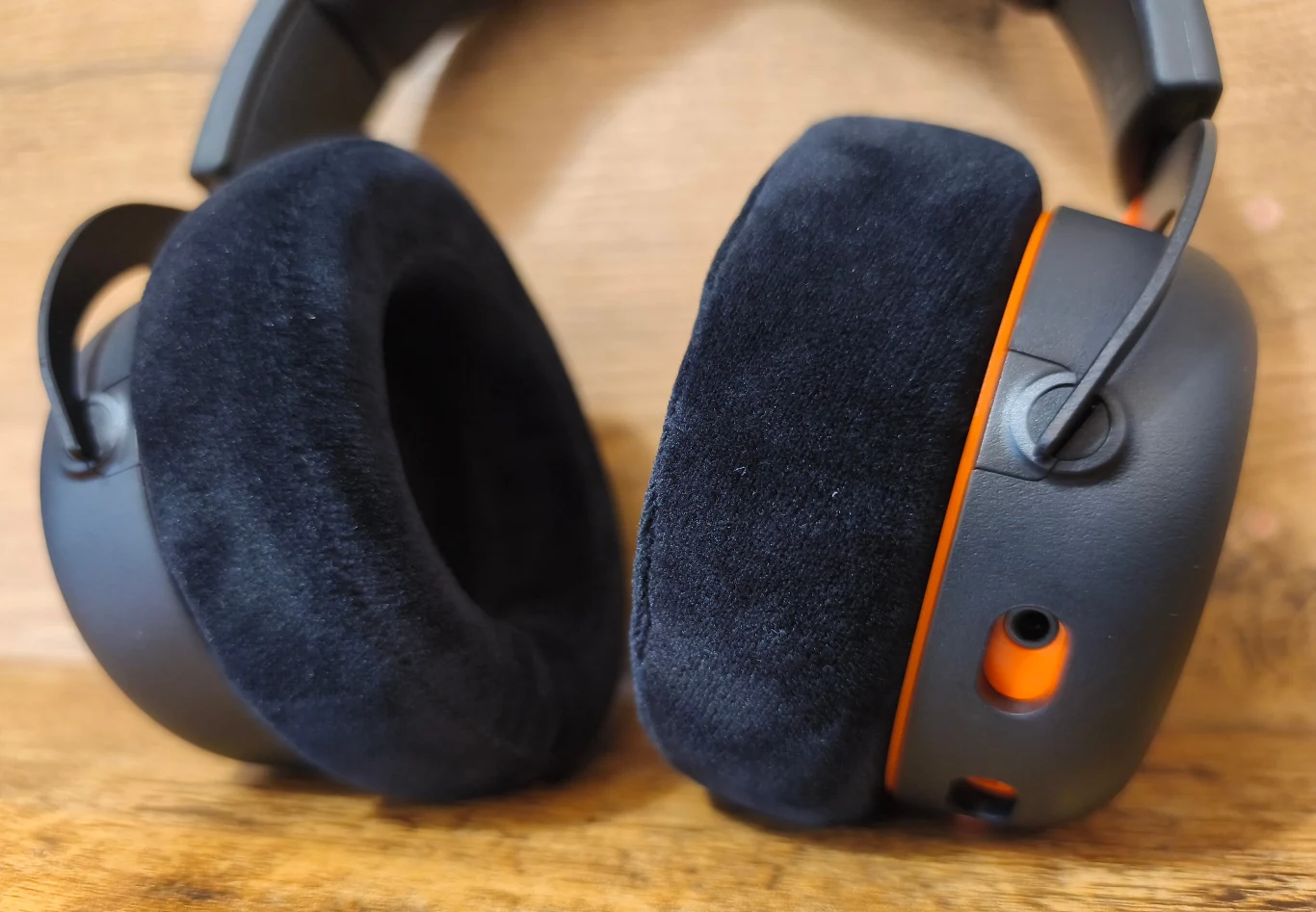 Beyerdynamic MMX 150 Wireless Duże nauszniki słuchawek z miękkimi, welurowymi poduszkami oraz widocznymi pomarańczowymi elementami na obudowie, całość na tle drewnianej powierzchni.