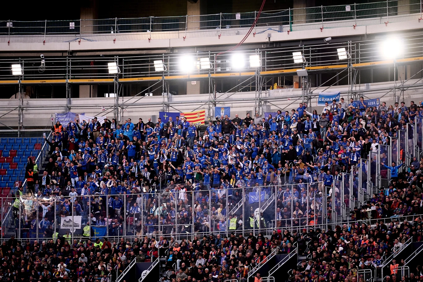 Kibice Realu Oviedo podczas meczu z Barceloną Tłum kibiców ubranych w niebieskie barwy zgromadzony na trybunach stadionu, oddzielony przezroczystą barierą ochronną, w tle widoczne światła stadionowe oraz fragmenty trybun z niewielką liczbą widzów.