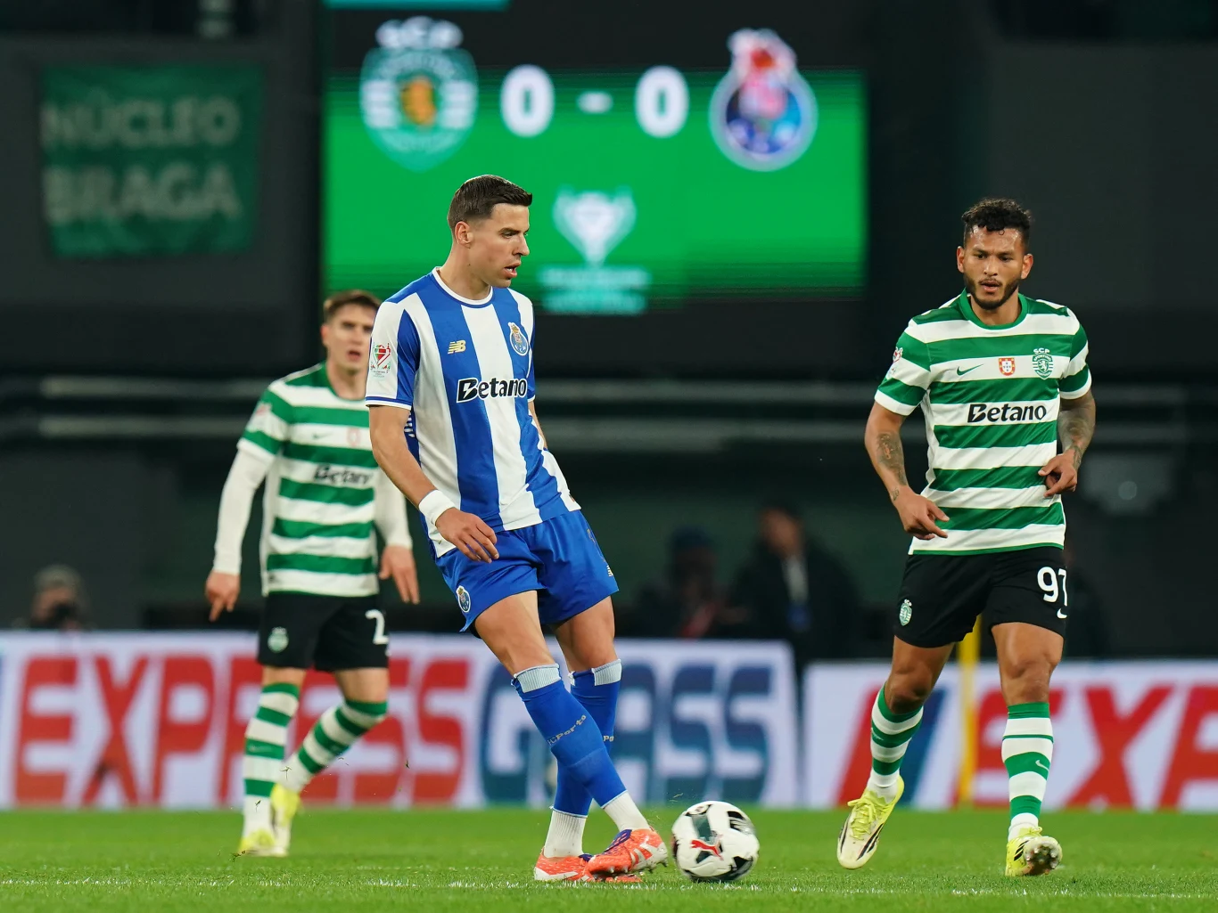 Piłkarz w niebiesko-białym stroju drybluje piłkę na boisku piłkarskim, otoczony przez dwóch zawodników w zielono-białych pasiastych koszulkach podczas meczu ligowego, na tle elektronicznej tablicy wyników pokazującej remis 0-0.