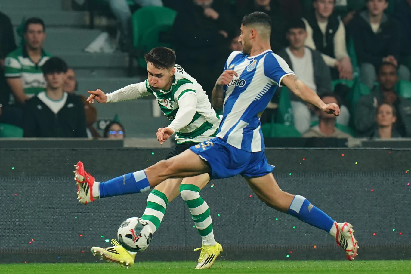 Dwóch piłkarzy rywalizujących o piłkę podczas meczu; zawodnik w zielono-białych pasach wykonuje dynamiczny zwód, podczas gdy przeciwnik w niebiesko-białym stroju próbuje zablokować jego ruch zdecydowanym wślizgiem.