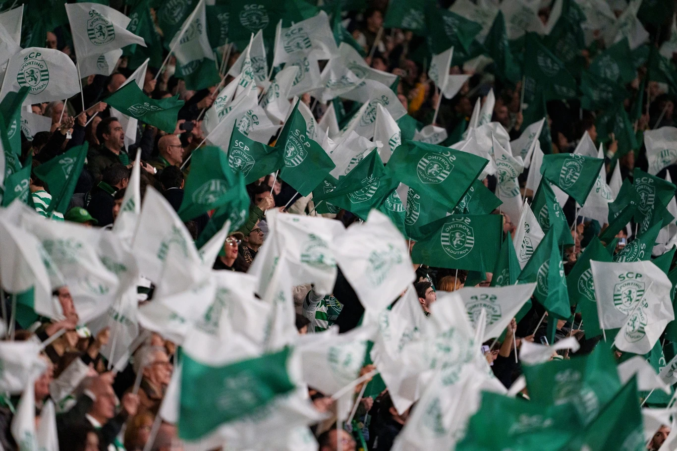 Tłum kibiców z zielono-białymi flagami z herbem klubu piłkarskiego, unoszącymi się w powietrzu, tworzących dynamiczną, pełną energii atmosferę stadionową.
