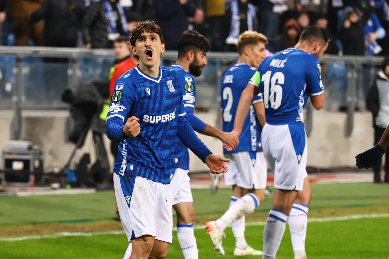 Piłkarz w niebieskim stroju wyraża silne emocje na boisku, zaciskając pięść i otwierając usta w geście triumfu, wokół niego kilku innych zawodników w podobnych strojach, skupionych na grze.