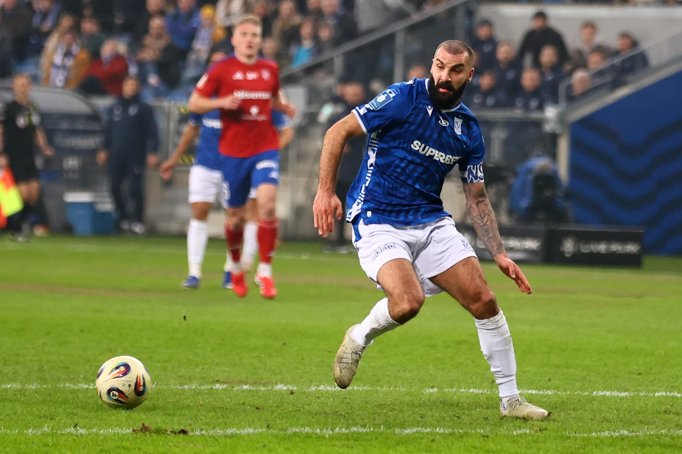 Mikael Ishak piłkarz w niebieskiej koszulce i białych spodenkach kontroluje piłkę na murawie podczas meczu, skupiony na grze; w tle inni zawodnicy oraz kibice na trybunach