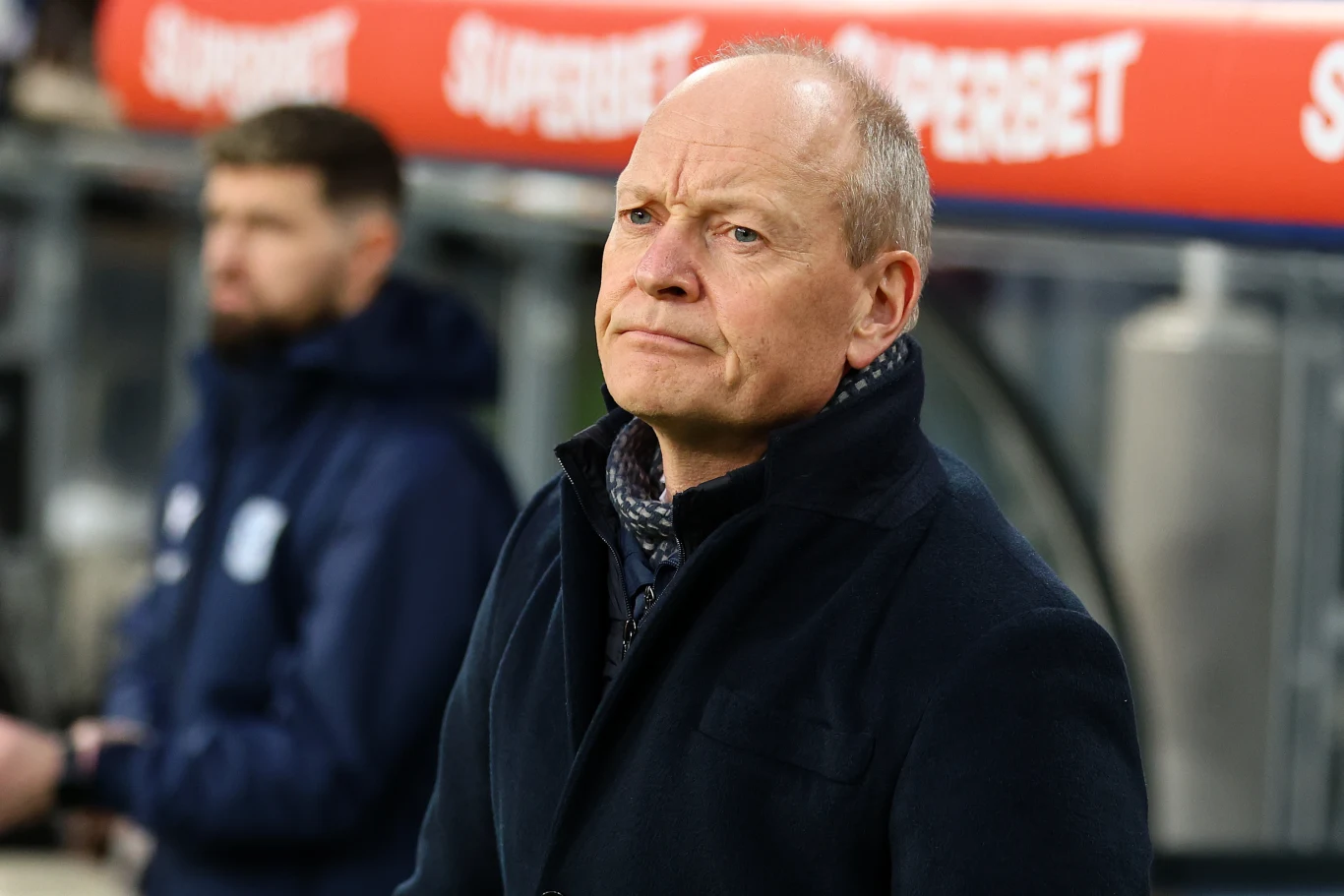 Niels Frederiksen, trener Lecha Poznań Mężczyzna w średnim wieku w ciemnym płaszczu stoi na tle stadionu piłkarskiego, z poważnym wyrazem twarzy; w tle widoczna rozmazana postać drugiego mężczyzny oraz fragmenty banera reklamowego.