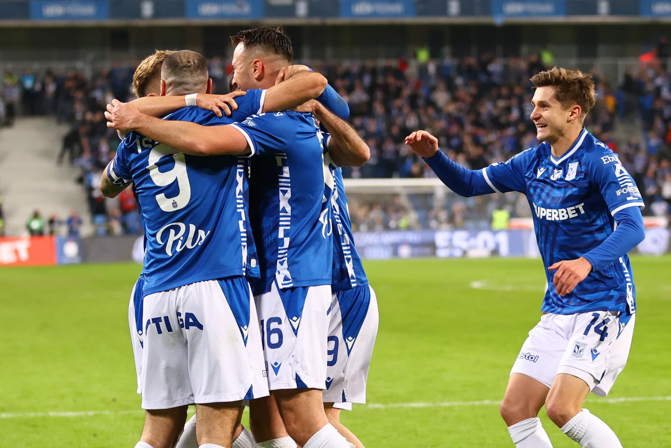 Lech Poznań wygrał z Rakowem Częstochowa Piłkarze w niebieskich strojach celebrują zdobycie gola na boisku, obejmując się i wyrażając radość, podczas gdy jeden z zawodników podnosi rękę w geście triumfu.