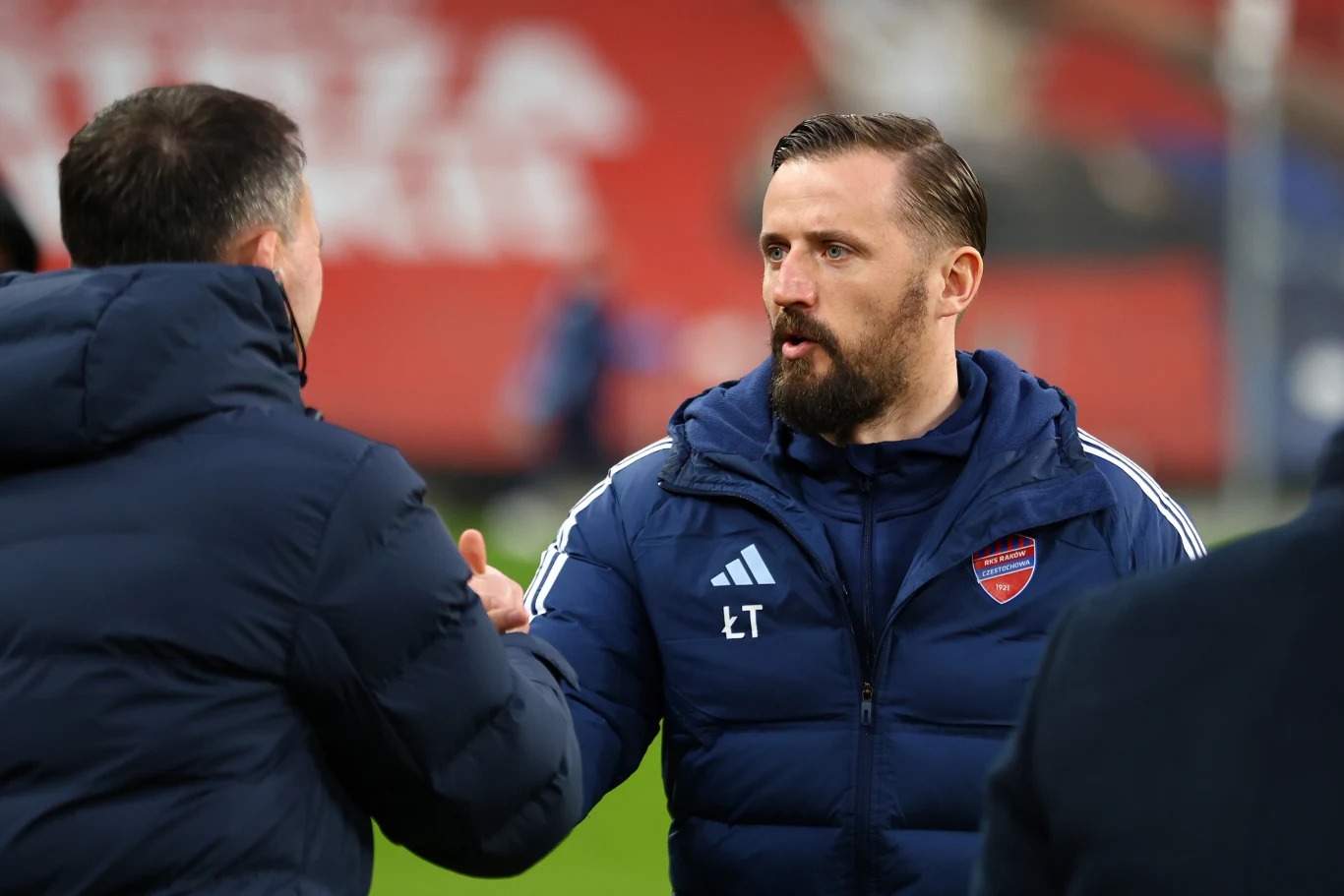 Mężczyzna w sportowej kurtce z logo klubu piłkarskiego i inicjałami 'LT' podaje rękę innemu mężczyźnie w kurtce, prowadząc rozmowę na tle stadionu sportowego.