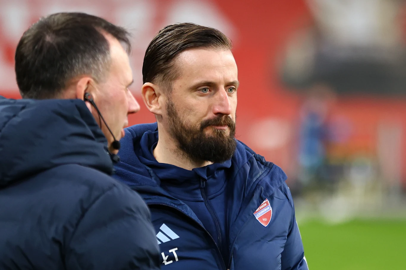 Dwóch mężczyzn w sportowych kurtkach rozmawia na tle rozmytego stadionu; jeden z nich ma brodę i herb klubu sportowego na ubraniu.