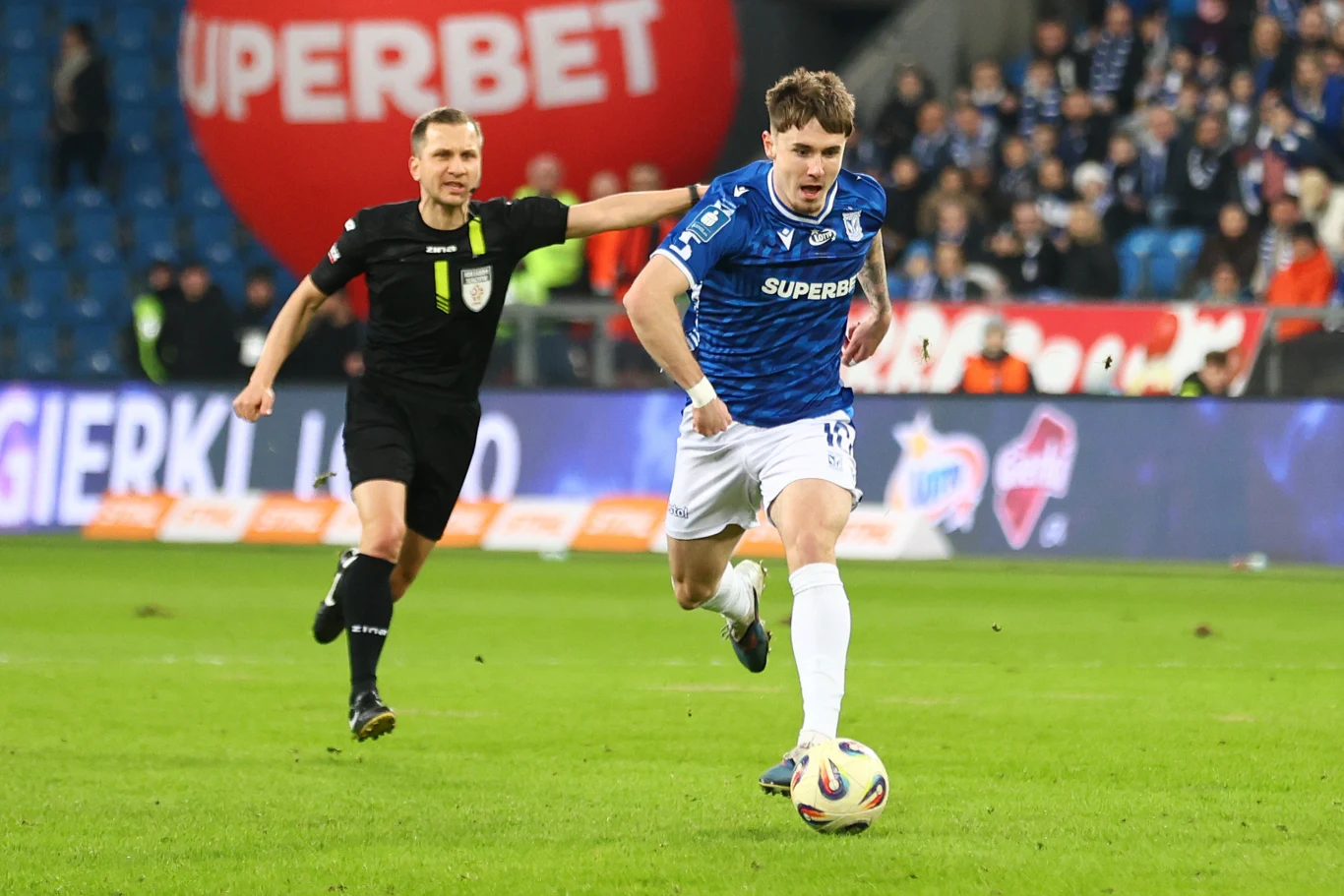 Zawodnik w niebieskiej koszulce biegnie z piłką po murawie stadionu, podczas gdy sędzia zbliża się z tyłu; w tle widoczni kibice i duży balon z logo sponsora.