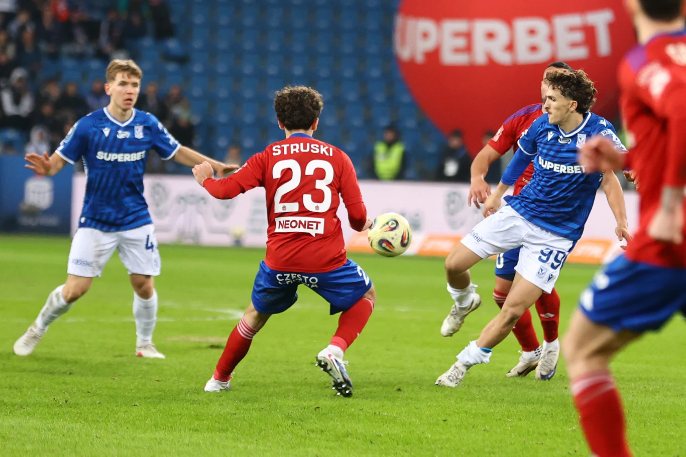 Dwóch piłkarzy w niebieskich koszulkach walczy o piłkę z zawodnikiem w czerwonym stroju, który ustawia się do odbioru futbolówki na środku boiska podczas meczu ligowego, w tle widoczna pusta trybuna oraz duży balon z napisem sponsorującym.