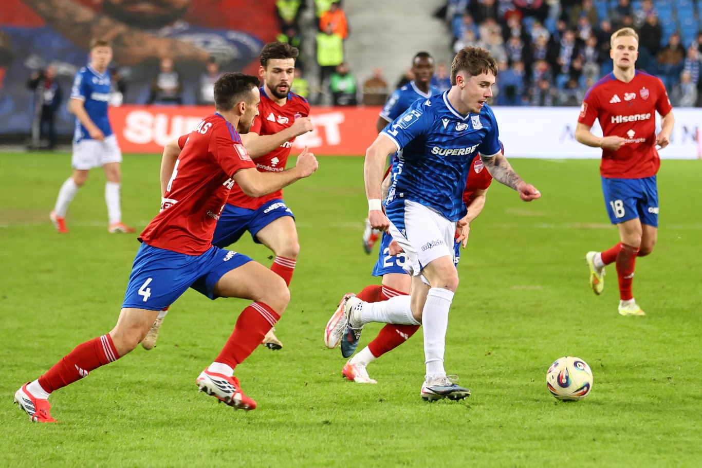 Piłkarz w niebieskiej koszulce biegnie z piłką, ścigany przez zawodników w czerwonych strojach na tle stadionu pełnego kibiców.