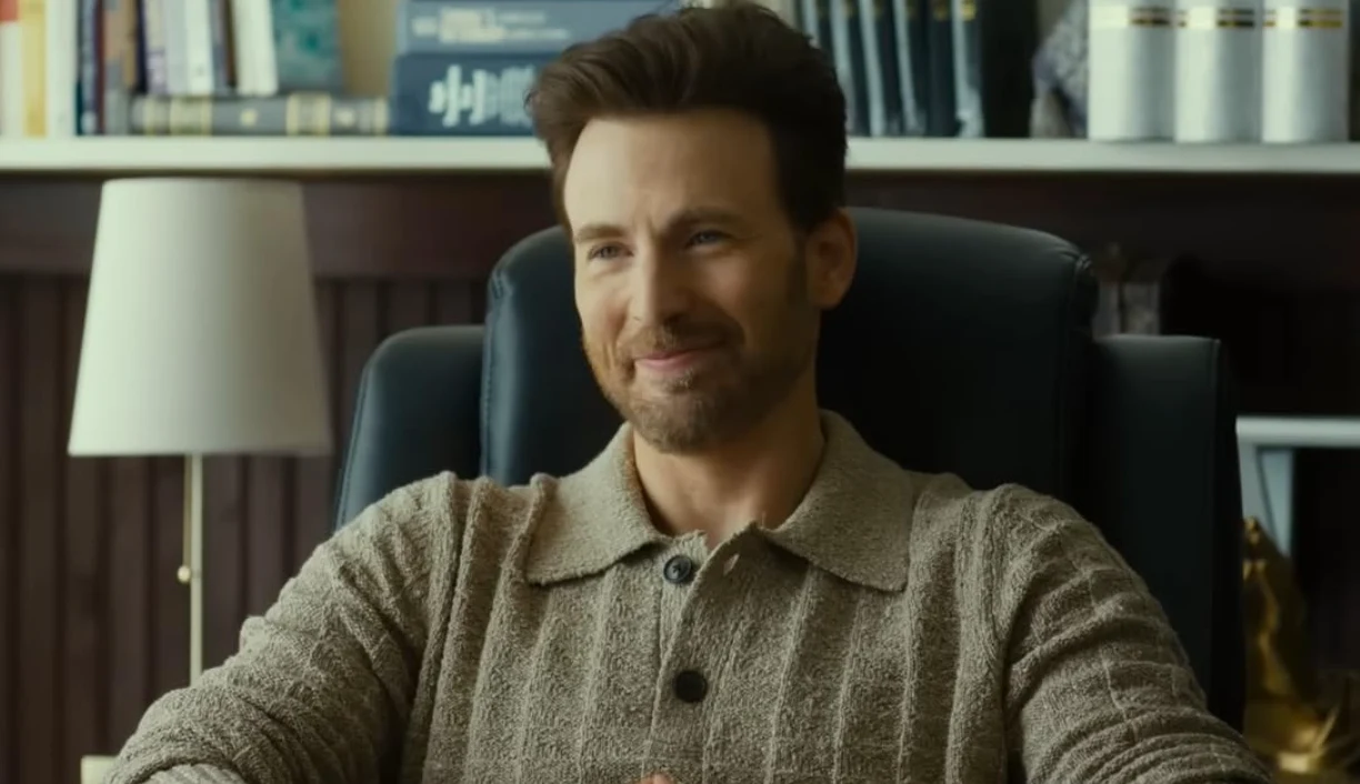 Uśmiechnięty mężczyzna z przyciemnionym zarostem siedzi na czarnym fotelu biurowym, ubrany w beżowy sweter, w tle regał z książkami i lampa stojąca na stole.