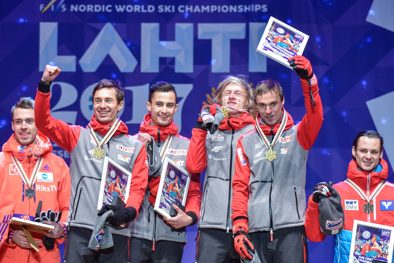 Grupa uśmiechniętych sportowców ubranych w zimowe kurtki i bluzy, stojąca na podium z medalami i dyplomami, unosząca w geście zwycięstwa ręce, na tle niebieskiej planszy z napisem 'LAHTI 2017' podczas ceremonii wręczania nagród na mistrzostwach narciar...