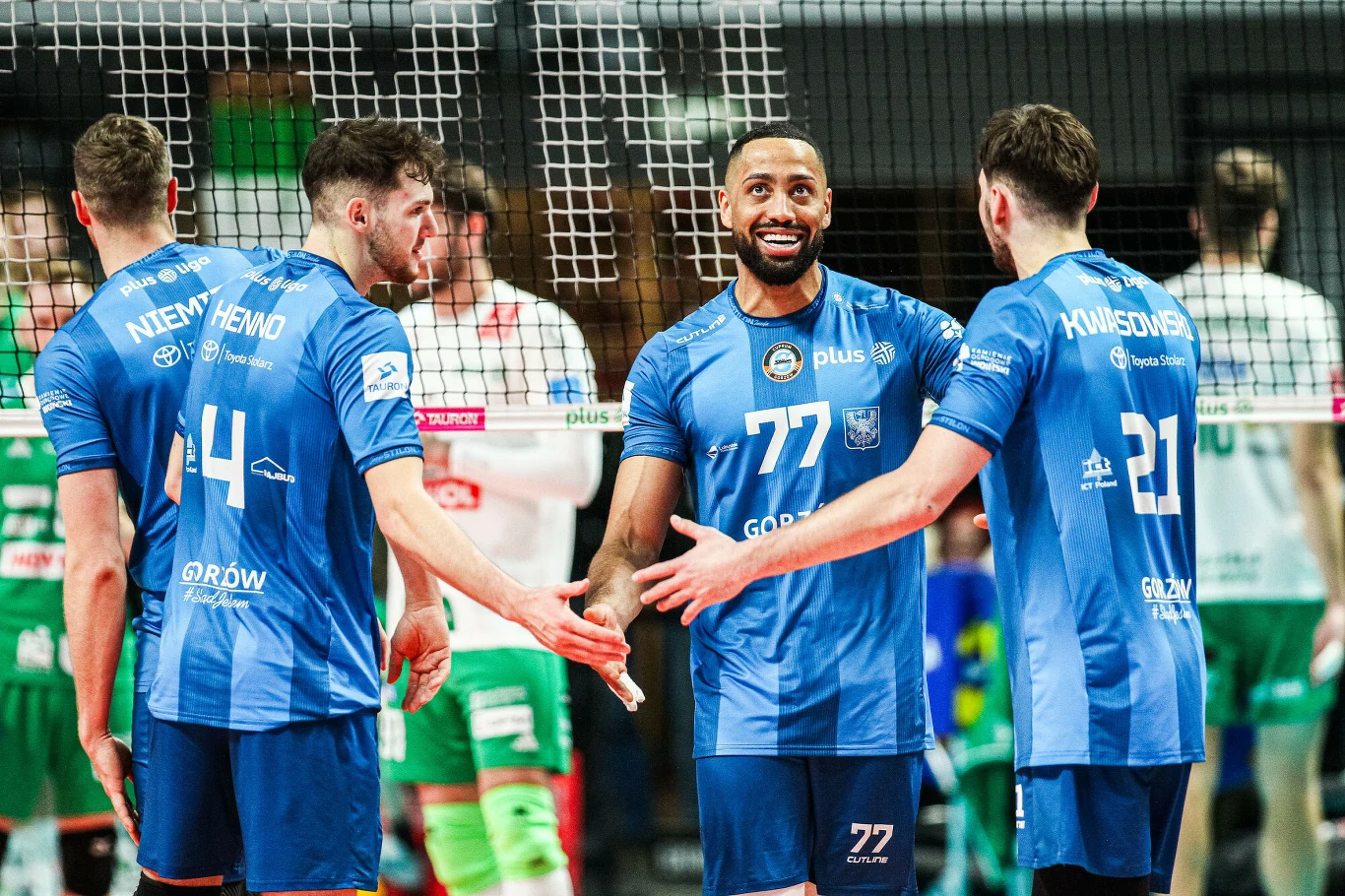 Trzech siatkarzy drużyny w niebieskich koszulkach cieszy się i przybija piątki po udanej akcji, na twarzach widać uśmiechy, w tle siatka i zawodnicy przeciwnej drużyny w zielono-białych strojach.