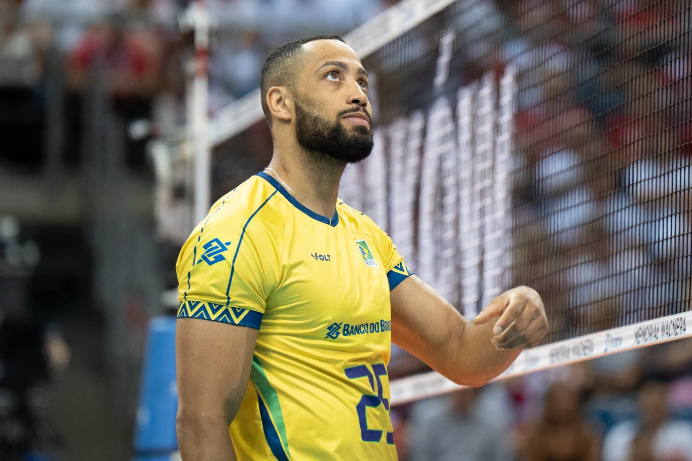 Siatkarz w żółtej koszulce z numerem 23 stoi przy siatce na hali sportowej, skupiony, z ręką opartą na taśmie siatki, w tle widoczni kibice na trybunach.