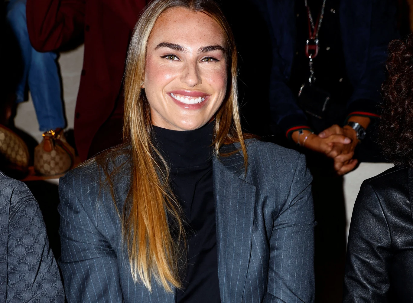 Aryna Sabalenka Aryna Sabalenka w szarym garniturze w prążki i czarnym golfie, siedzi na tle ciemnych sylwetek.