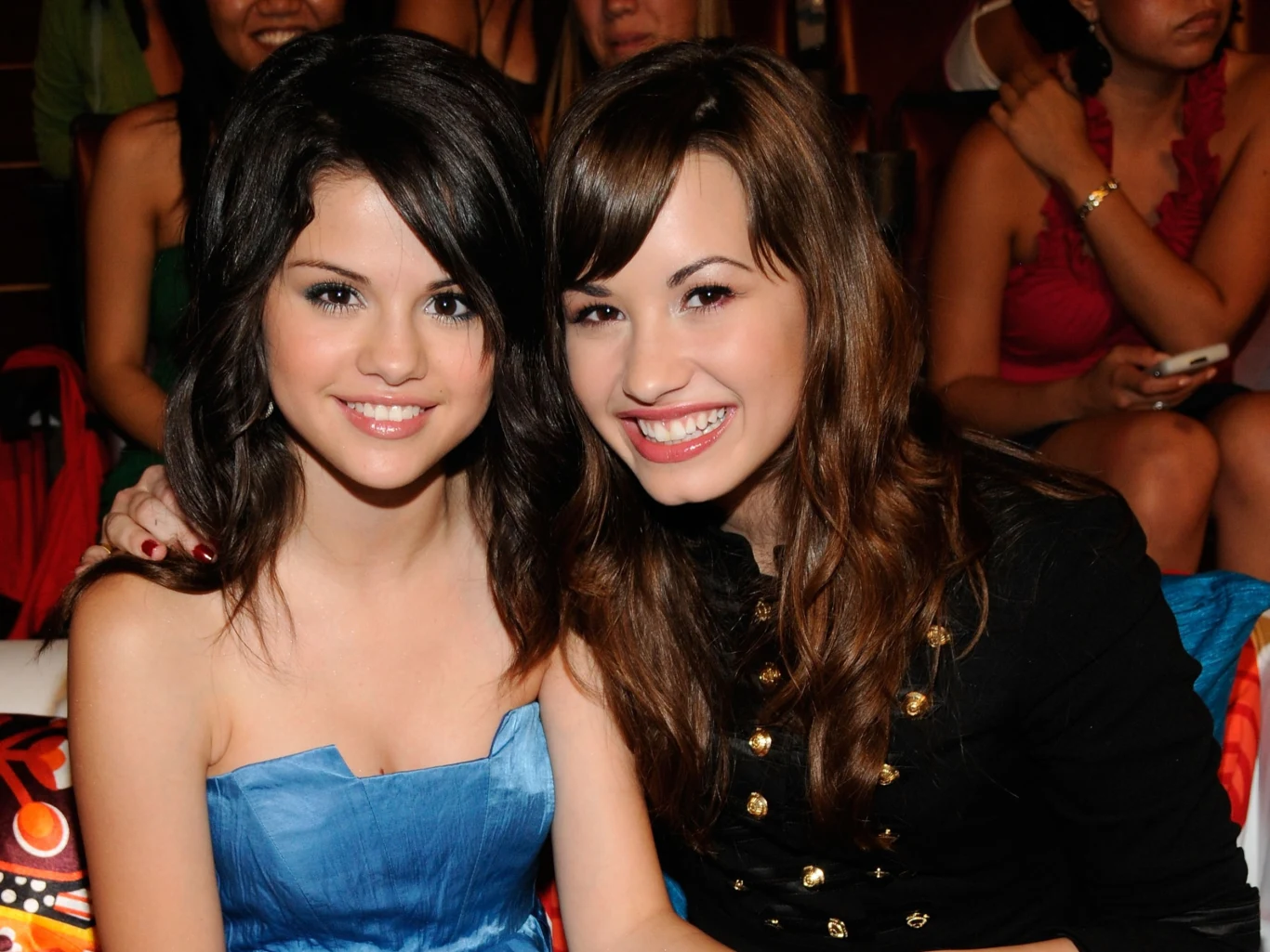 Dwie młode kobiety siedzą obok siebie i uśmiechają się do aparatu; jedna ma na sobie niebieską sukienkę, druga czarną marynarkę z ozdobnymi guzikami, a obie mają ciemne, rozpuszczone włosy.