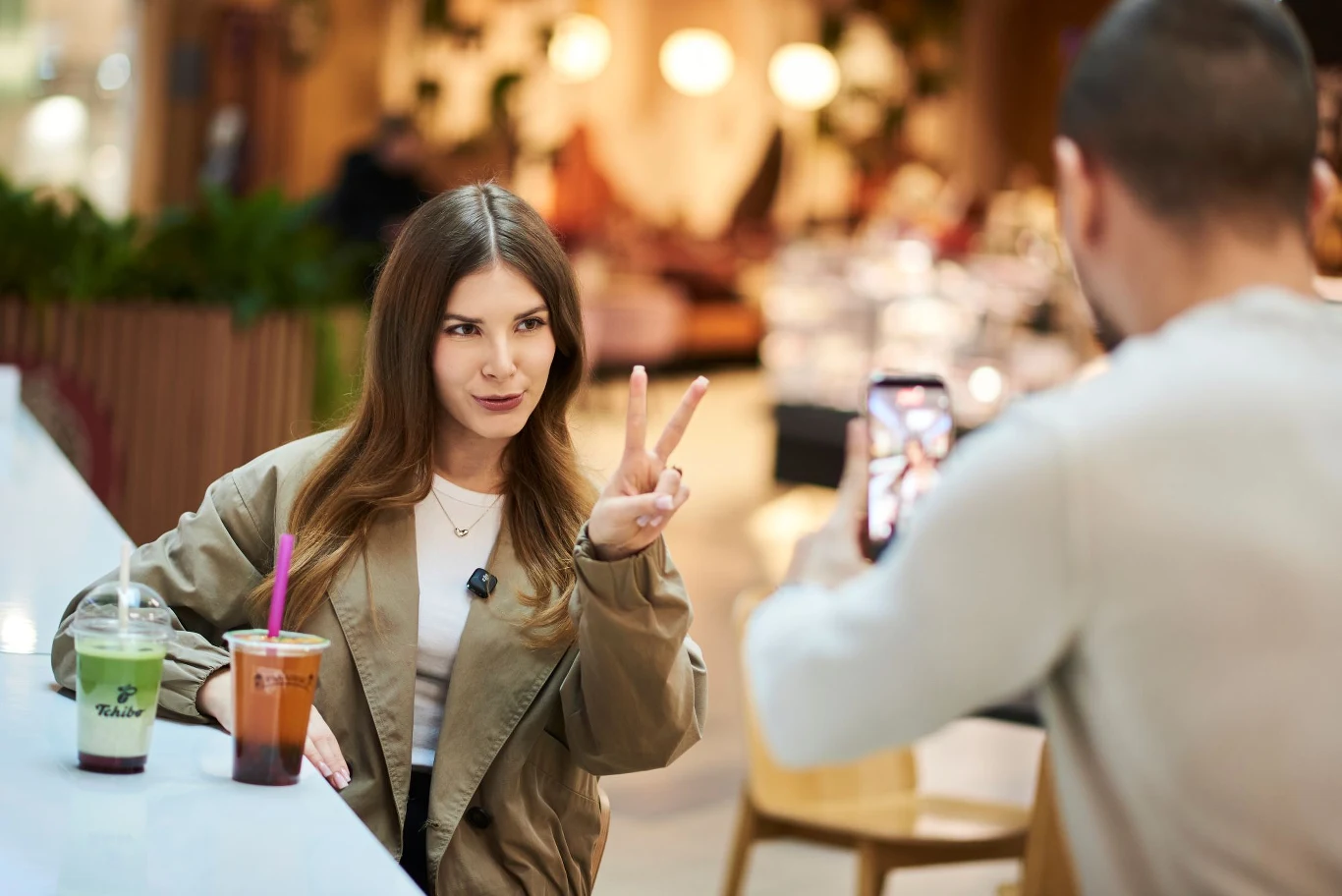 Młoda kobieta siedzi przy stole w kawiarni, pokazuje znak pokoju do aparatu trzymanego przez drugą osobę, przed nią stoją dwa napoje z plastikowymi słomkami.