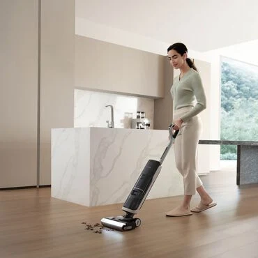 Kobieta sprząta drewnianą podłogę w nowoczesnej kuchni przy użyciu bezprzewodowego odkurzacza pionowego, usuwając rozsypane drobne resztki.