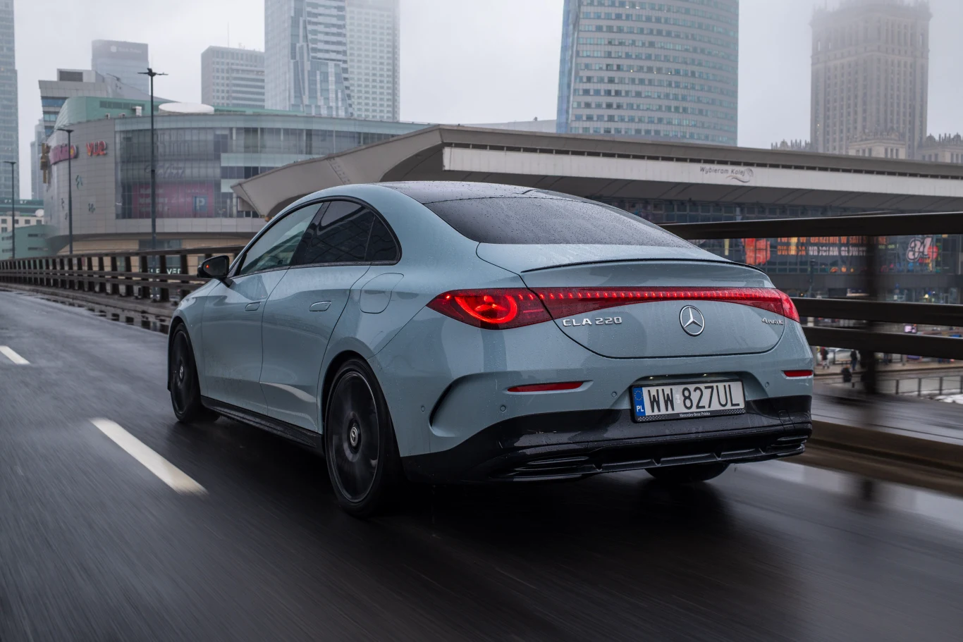 Największa pojedyncza zaleta? Doskonały komfort jazdy, niezależnie od tego, co ma pod kołami. Jasnoniebieski Mercedes-Benz EQE 350 jadący po miejskiej estakadzie, otoczony nowoczesnymi wieżowcami i charakterystyczną miejską architekturą w tle przy pochmurnej pogodzie.