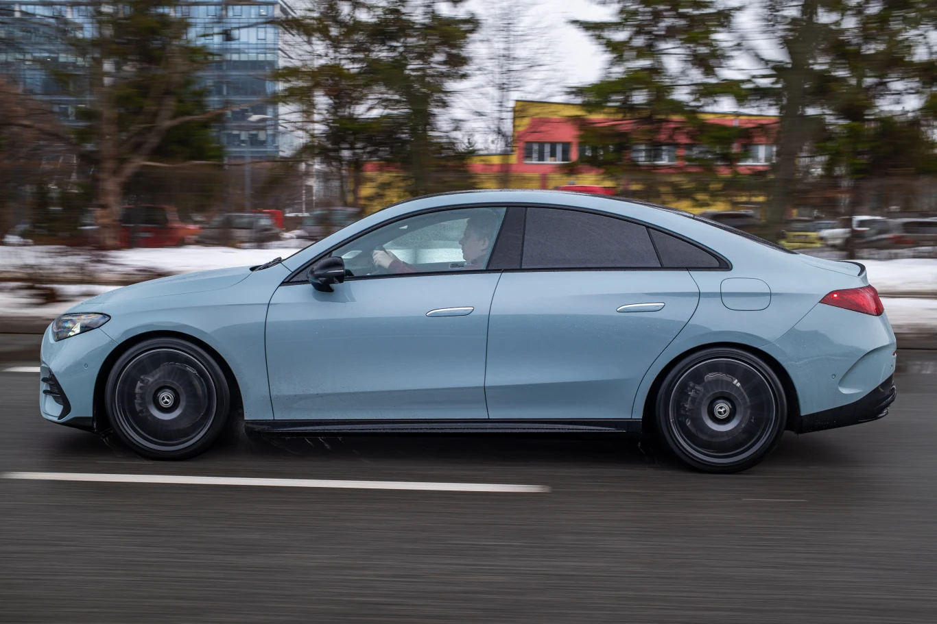 Nowe CLA jest dłuższe od poprzednika o 3,5 cm - efekt tego czuć w kabinie. Jasnoniebieski nowoczesny sedan w ruchu na miejskiej drodze, z czarnymi felgami i przyciemnianymi szybami, na tle zimowego krajobrazu z drzewami i śniegiem po bokach jezdni.