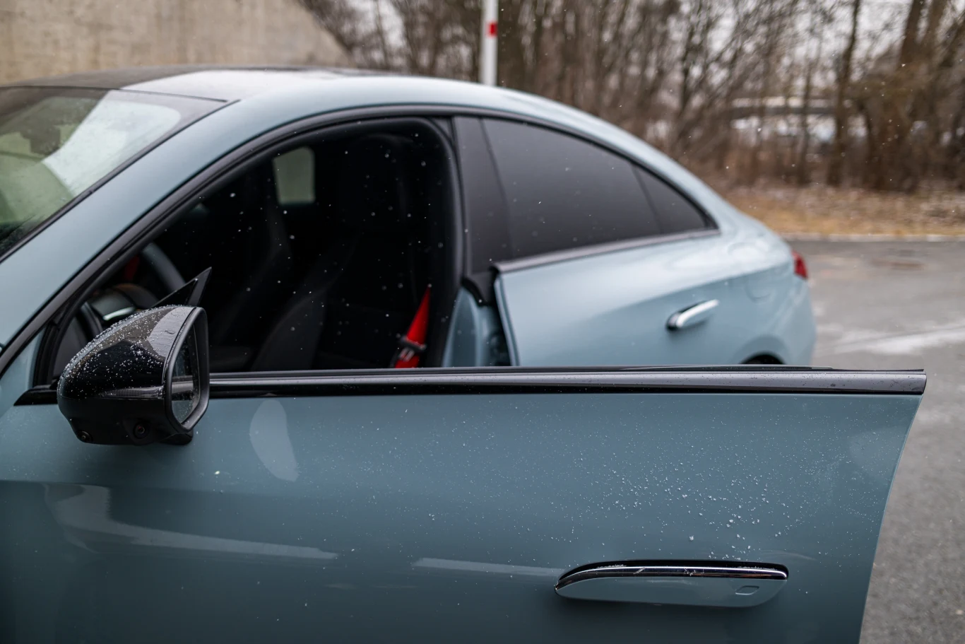 Szyby bez ramek nie wpływają na poziom hałasu we wnętrzu nawet przy prędkościach autostradowych. Jasnoniebieski samochód z otwartymi przednimi drzwiami na tle betonowego muru i bezlistnych drzew, widoczne czarne przyciemniane szyby oraz lusterko boczne pokryte drobnymi kroplami wody.