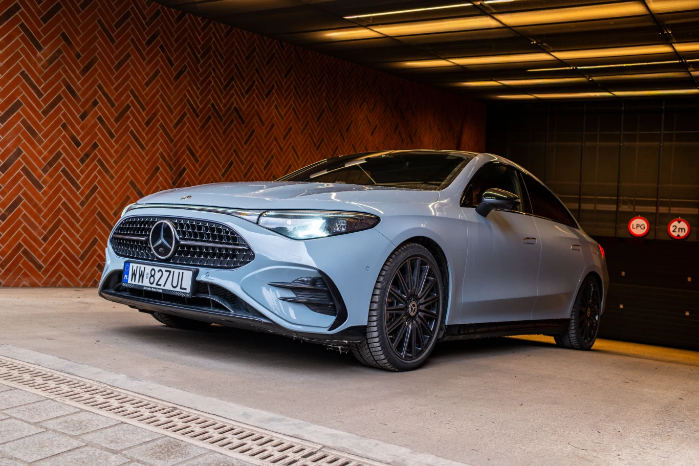 Połyskujące czernią elementy to część pakietu AMG Line Plus za ponad 24 tys. zł. Srebrny Mercedes klasy A z czarnymi felgami stoi na podjeździe przy czerwonej ceglanej ścianie, na tle oświetlonego sufitu i zamkniętej bramy z ostrzegawczymi znakami o ograniczeniu prędkości i wysokości.