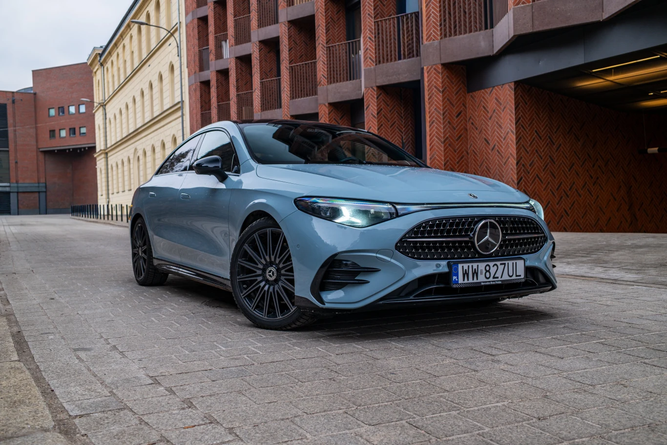 Od wersji elektrycznej hybrydowe CLA różni się przede wszystkim klasyczną osłoną chłodnicy - choć nadal jej wzór składa się z małych gwiazdek. Jasnoniebieski Mercedes klasy A zaparkowany na brukowanej ulicy między nowoczesnym i klasycznym budynkiem, z nowoczesnym grillem i czarnymi alufelgami na pierwszym planie.