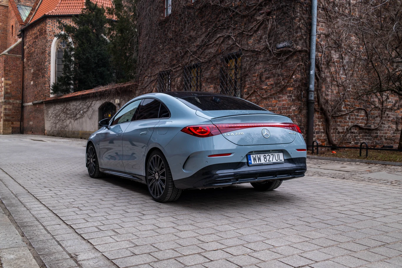 CLA wygląda nowocześnie, a szczególnie przyciąga wzrok wtedy, gdy robi się ciemno. Srebrny Mercedes-Benz CLA stoi zaparkowany na brukowanej ulicy, otoczony przez zabytkowe mury z cegły porośnięte bluszczem.