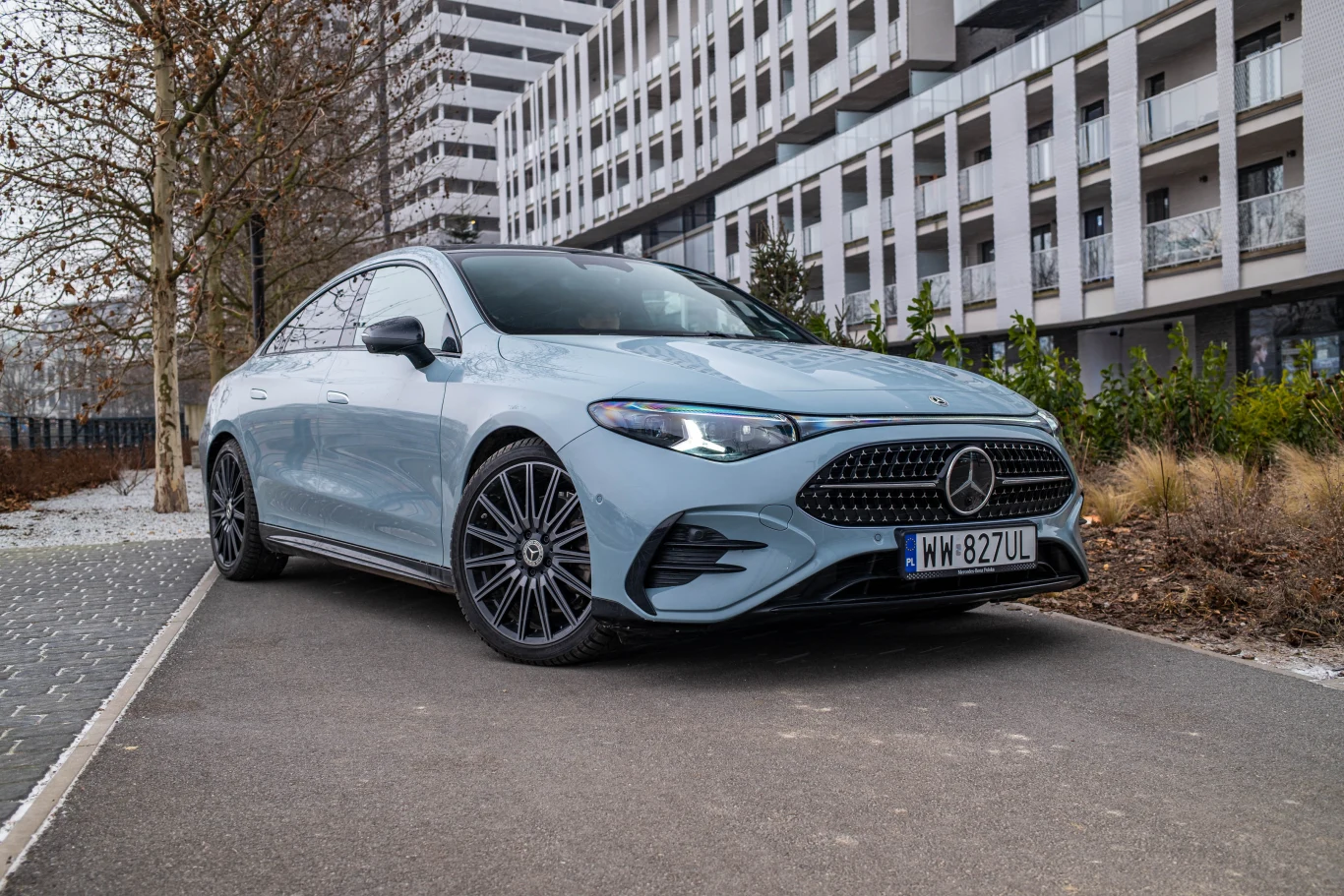 Gwiazda to motyw przewodni nadwozia - takim wzorem świecą się między innymi światła do jazdy dziennej. Jasnoniebieski Mercedes-Benz zaparkowany na ścieżce wśród nowoczesnych budynków mieszkalnych, otoczony zielenią oraz suchymi trawami, z widocznymi czarnymi felgami i charakterystycznym, sportowym grillem.