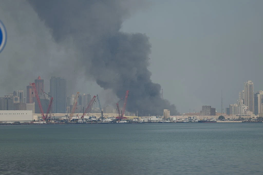 Gęsty czarny dym unosi się nad obszarem portowym miasta, w tle wysokie budynki i dźwigi portowe nad linią wody.
