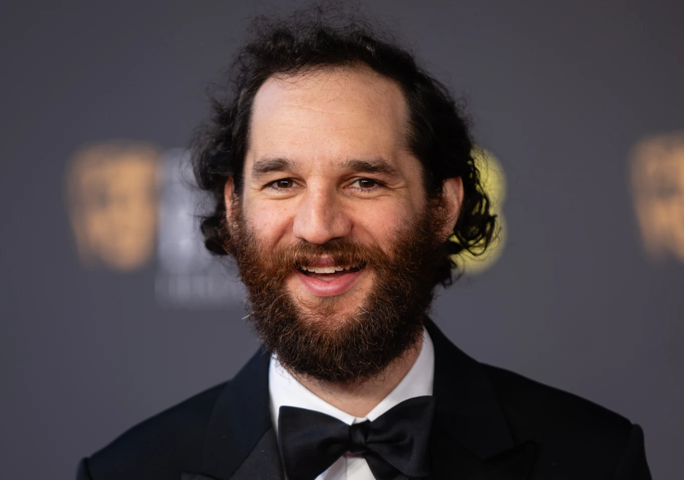 Josh Safdie na gali rozdania nagród BAFTA w 2026 roku Mężczyzna z gęstą brodą i kręconymi włosami, ubrany w czarny smoking z muszką, uśmiecha się na tle ciemnej ścianki z rozmytymi złotymi logotypami.