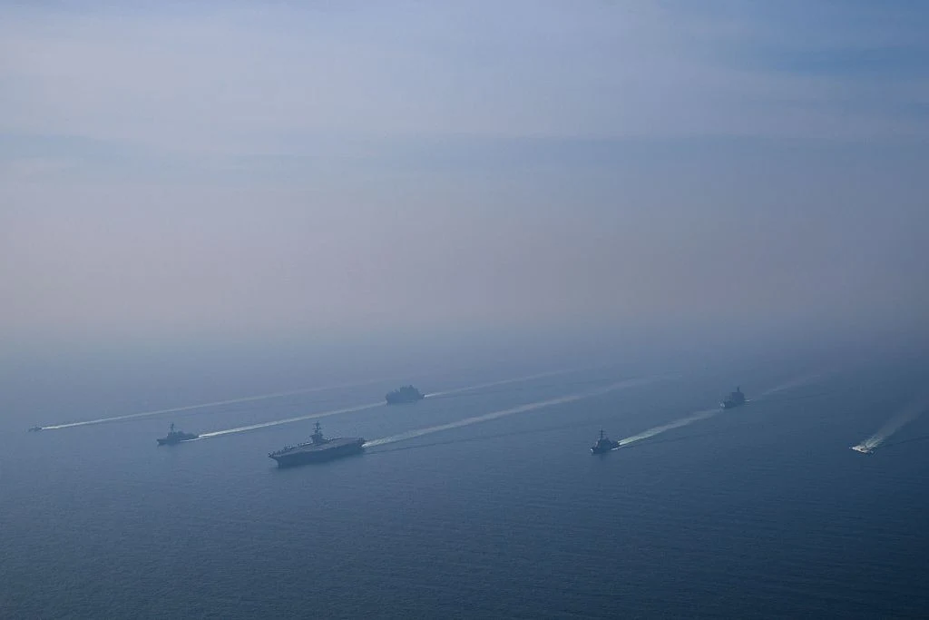 Formacja kilku dużych okrętów wojennych płynących równolegle po spokojnej, lekko zamglonej powierzchni morza, każdy z widoczną smugą wody za rufą.