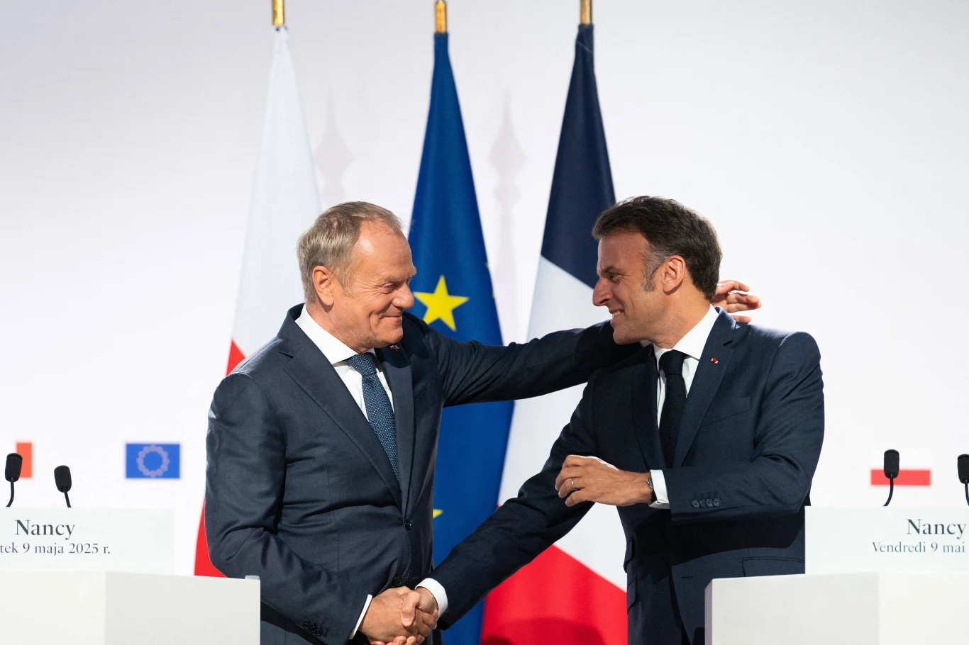 Donald Tusk i Emmanuel Macron podczas podpisywania polsko-francuskiej umowy w Nancy w 2025 roku Dwóch mężczyzn w garniturach podaje sobie ręce i uśmiecha się do siebie na tle flag Polski, Unii Europejskiej i Francji podczas oficjalnej konferencji.