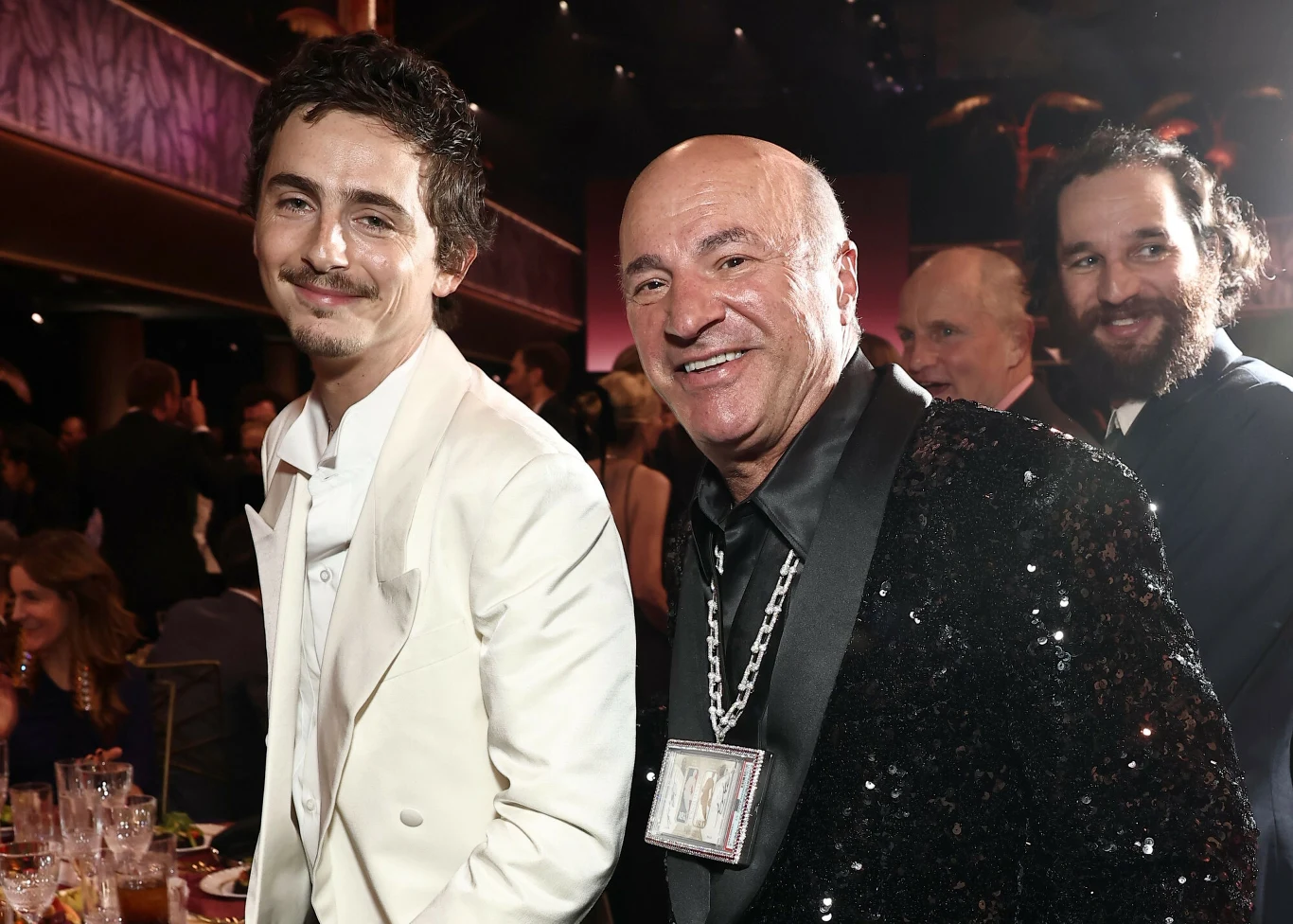 Timothée Chalamet, Kevin O'Leary i Josh Safdie na 32. gali rozdania nagród Aktorów Dwóch mężczyzn ubranych w eleganckie garnitury pozuje do zdjęcia podczas uroczystego wydarzenia, jeden z nich ma na sobie czarną marynarkę z cekinami oraz duży łańcuch na szyi, w tle widoczne są zastawione stoły oraz inni uczestnicy przyjęcia.