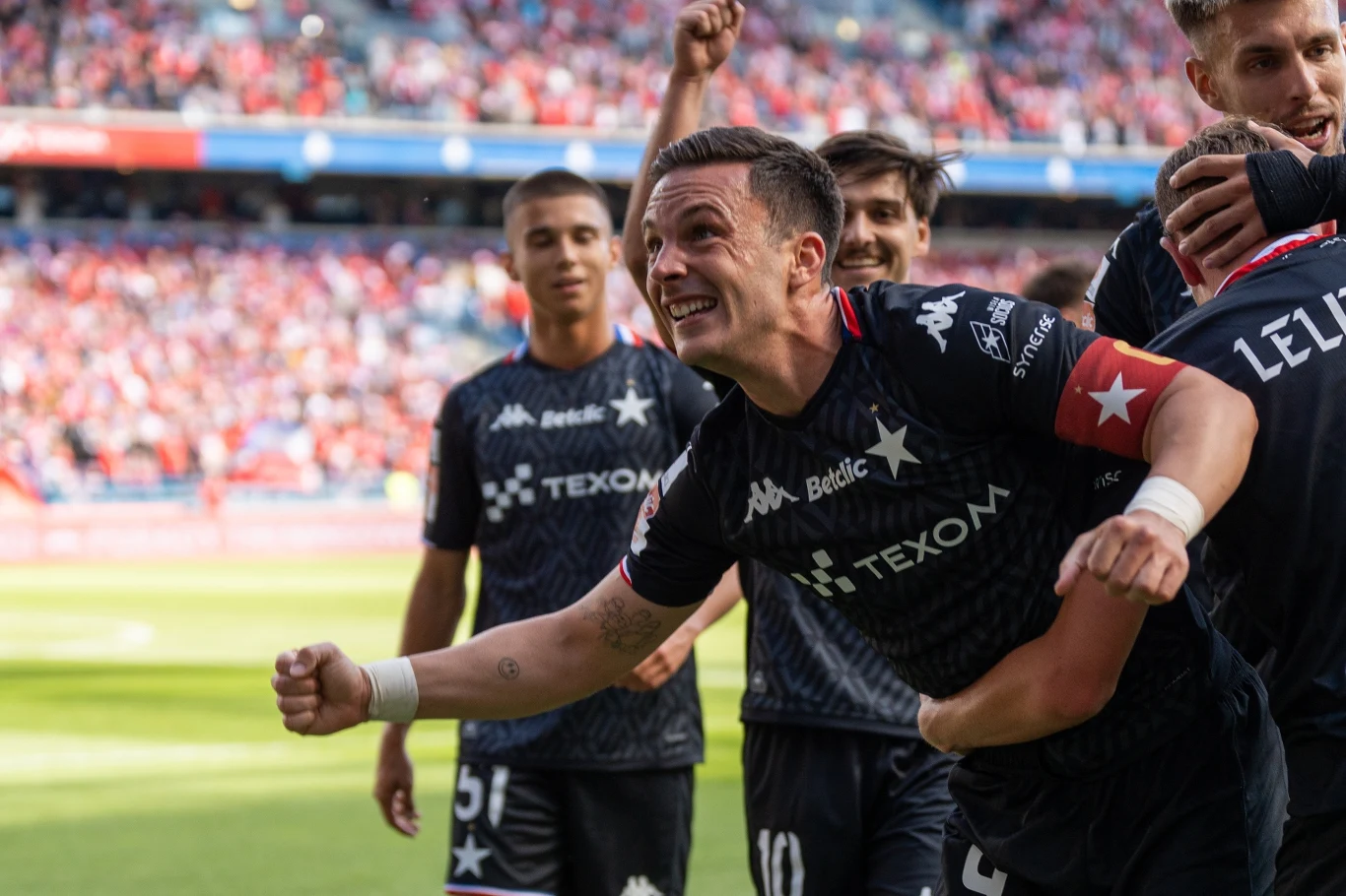 Piłkarz w czarnej koszulce z widoczną radością na twarzy celebruje zdobycie bramki wraz z kolegami z drużyny na stadionie pełnym kibiców.