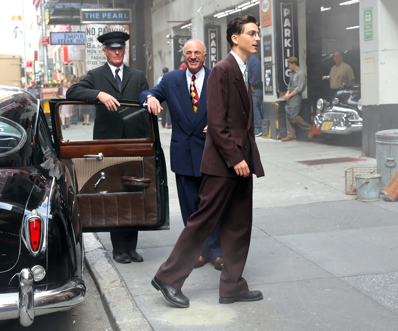 Kevin O'Leary i Timothee Chalamet na planie filmu "Wielki Marty" w październiku 2024 roku Mężczyzna w brązowym garniturze idzie chodnikiem obok eleganckiego czarnego samochodu z lat 50., podczas gdy elegancki szofer trzyma drzwi pojazdu, a uśmiechnięty starszy mężczyzna w kolorowym krawacie stoi w tle.