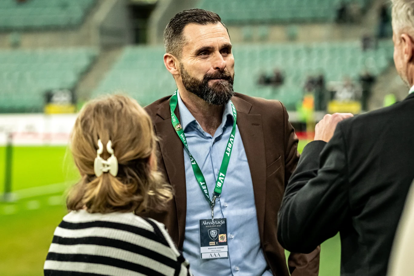 Mężczyzna z krótkim zarostem w brązowej marynarce i niebieskiej koszuli z identyfikatorem na szyi stoi na tle stadionu, rozmawiając z dwiema osobami, z których jedna ma związane włosy i nosi sweter w czarne pasy.