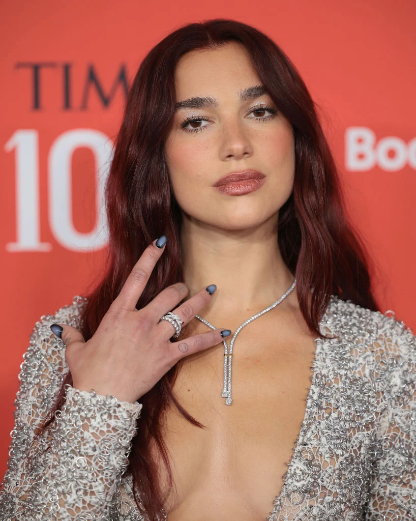 Kobieta o długich, ciemnorudych włosach, w srebrnej, błyszczącej sukni z głębokim dekoltem, prezentuje biżuterię: kilka pierścionków oraz elegancki naszyjnik na tle czerwonej ścianki z białymi napisami.