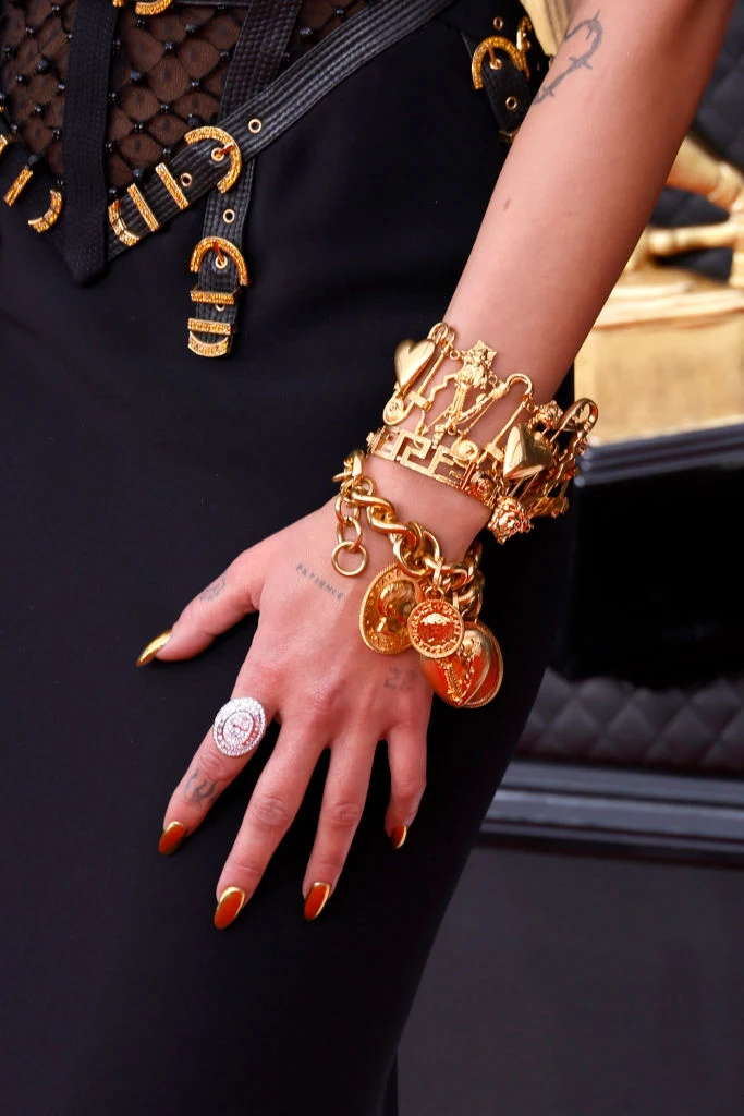 Dłoń ozdobiona licznymi złotymi bransoletkami z zawieszkami oraz medalionami, paznokcie pomalowane na złoty kolor, czarna sukienka z detalami z siatki i złotymi klamrami.