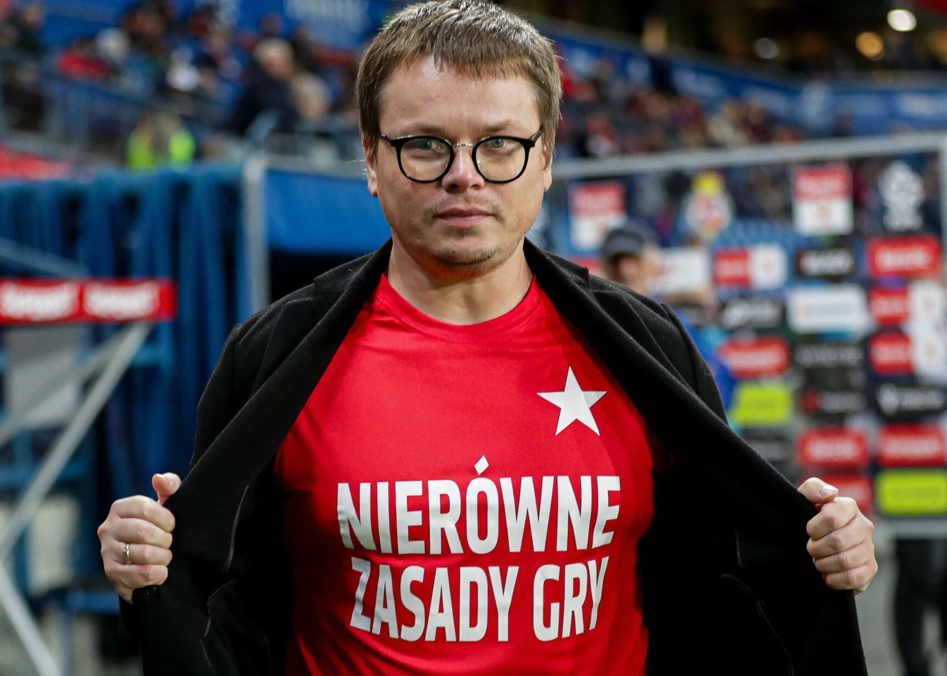 Mężczyzna w okularach i czarnej marynarce prezentuje czerwony t-shirt z napisem 'Nierówne zasady gry' oraz białą gwiazdą, stojąc na tle stadionu sportowego z widoczną tablicą reklamową.