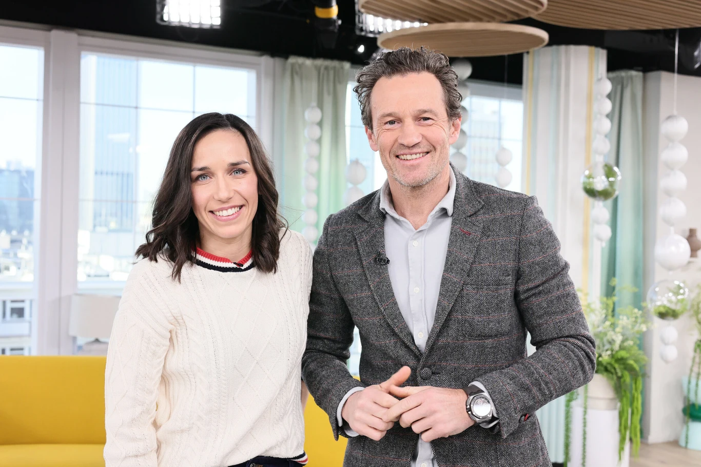 Kobieta w białym swetrze i mężczyzna w szarej marynarce stoją obok siebie z uśmiechami na twarzy w jasnym, nowoczesnym wnętrzu z żółtą sofą i dużymi oknami w tle.