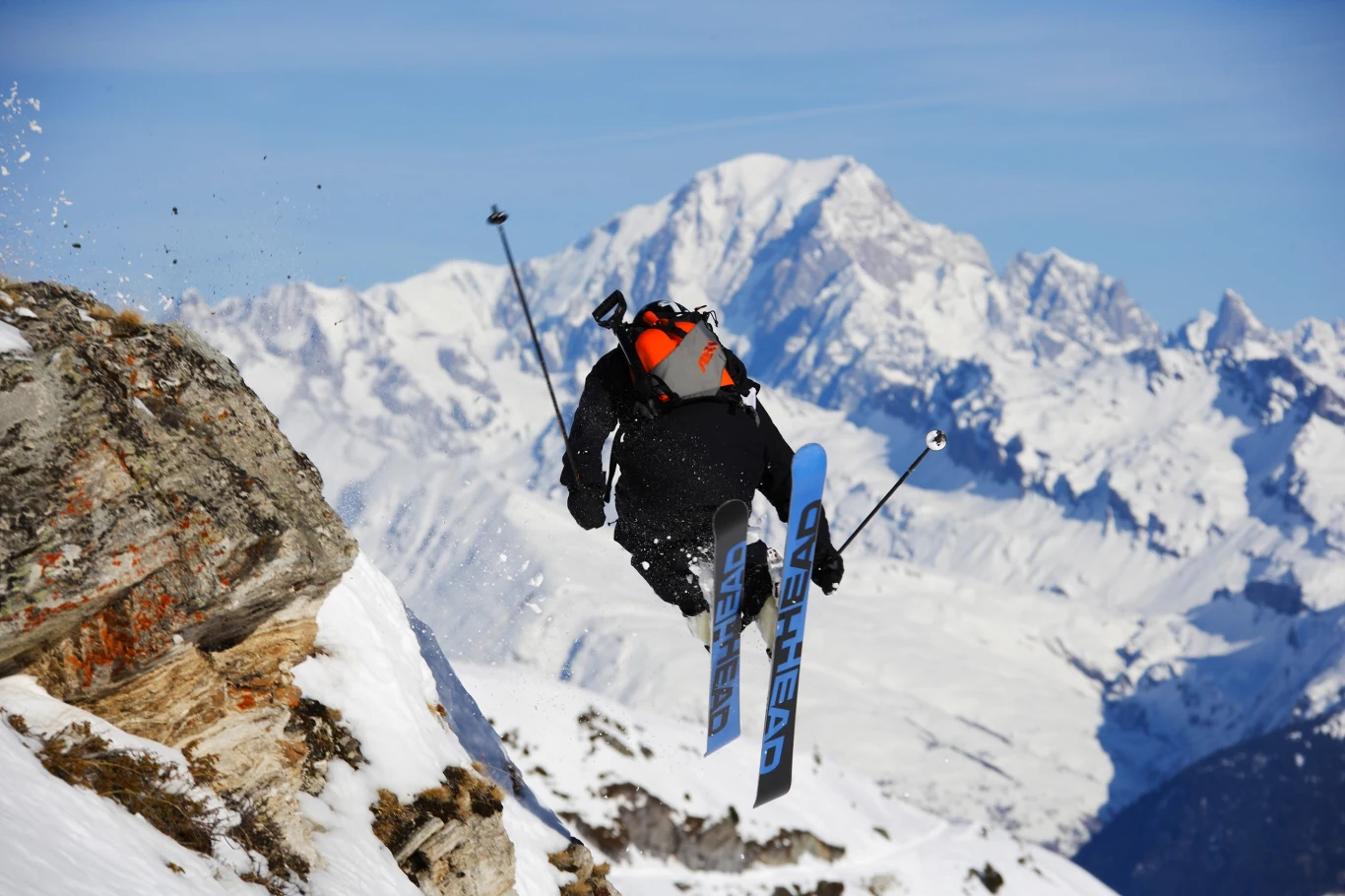 Narciarz wykonujący skok nad urwiskiem na tle wysokich, ośnieżonych gór; dynamiczna poza w powietrzu podkreśla sportowy charakter sceny.