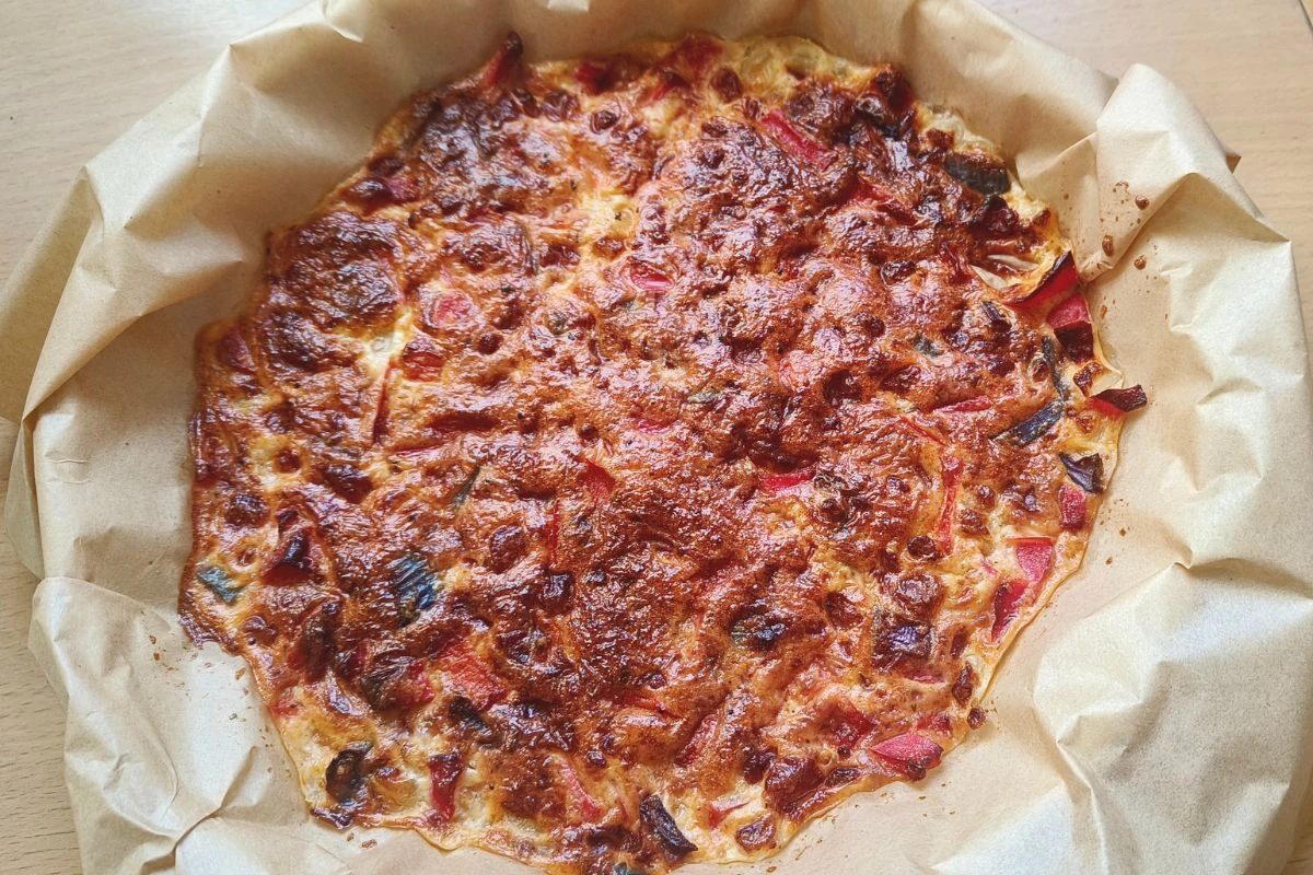 Duża, rumiana tarta wyłożona na papierze do pieczenia, z wyraźnie zarumienioną, serową górą oraz widocznymi kawałkami kolorowej papryki i ziół.