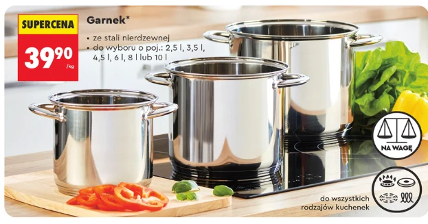 Garnki ze stali nierdzewnej sprzedawane na wagę 39,90 zł/kg – różne pojemności od 2,5 l do 10 l, źródło: gazetka Biedronka, str. 15 Trzy garnki ze stali nierdzewnej o różnych pojemnościach ustawione na kuchence indukcyjnej, obok pokrojone papryka i pietruszka, z tyłu świeże warzywa liściaste i żółta papryka, obok widoczne informacje o cenie i dostępnych opcjach pojemnościowych.