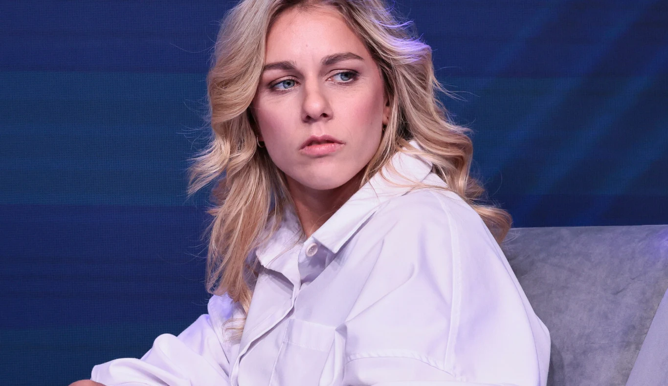 Aleksandra Mirosław Aleksandra Mirosław siedzi w białej koszuli na szarym fotelu, na tle ciemnoniebieskiej ściany.